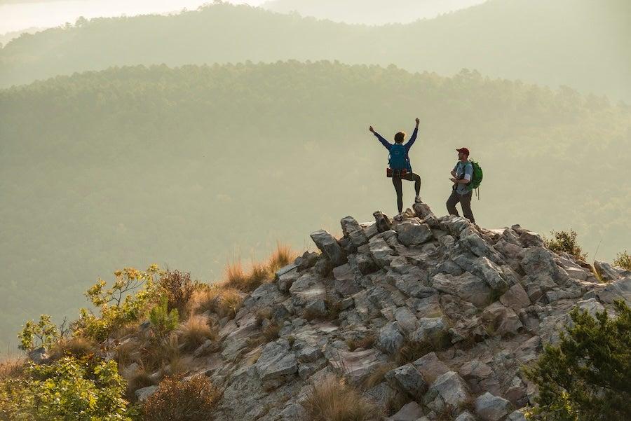 Two hikers celebrate on a rocky hilltop with misty mountains in the background.