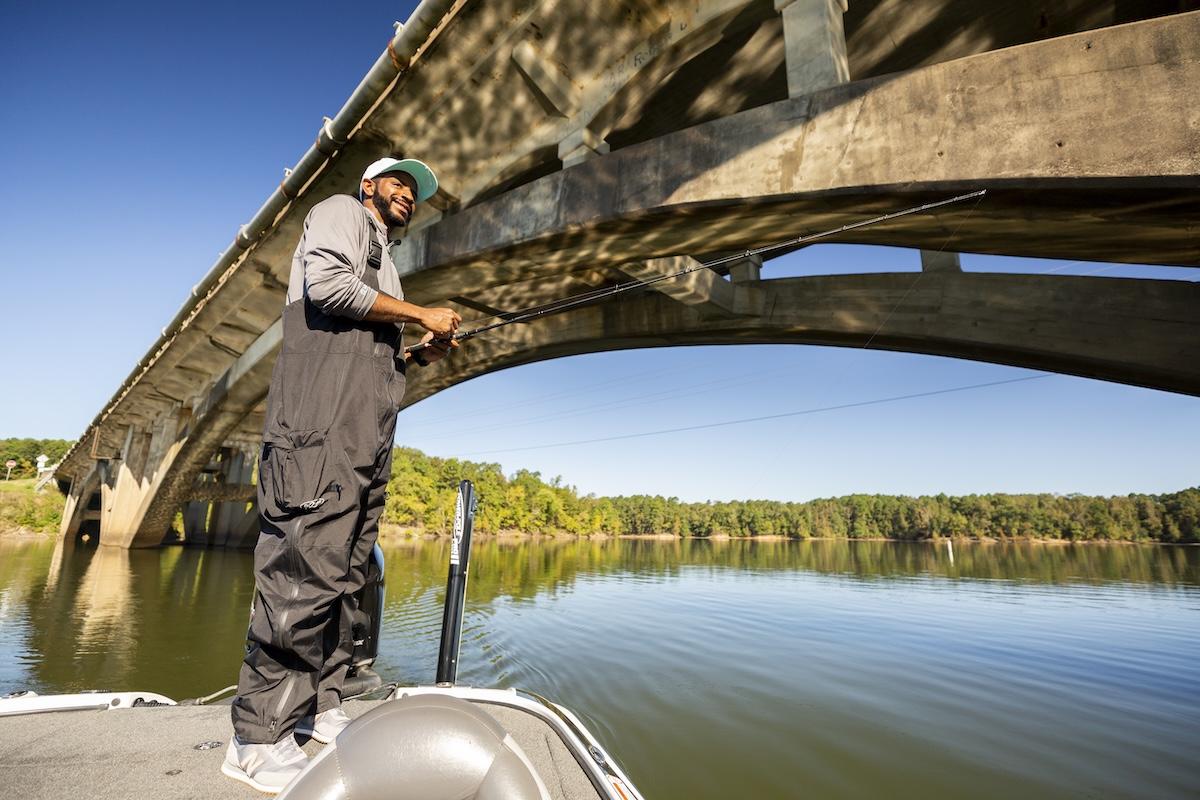 Man fishing from a boat under a large bridge on a sunny day.