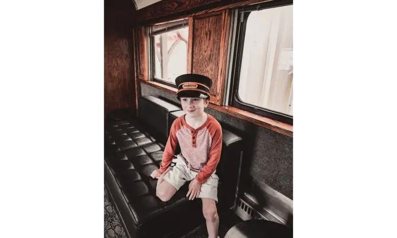 Child in conductor hat sitting in vintage train car with wooden interior.
