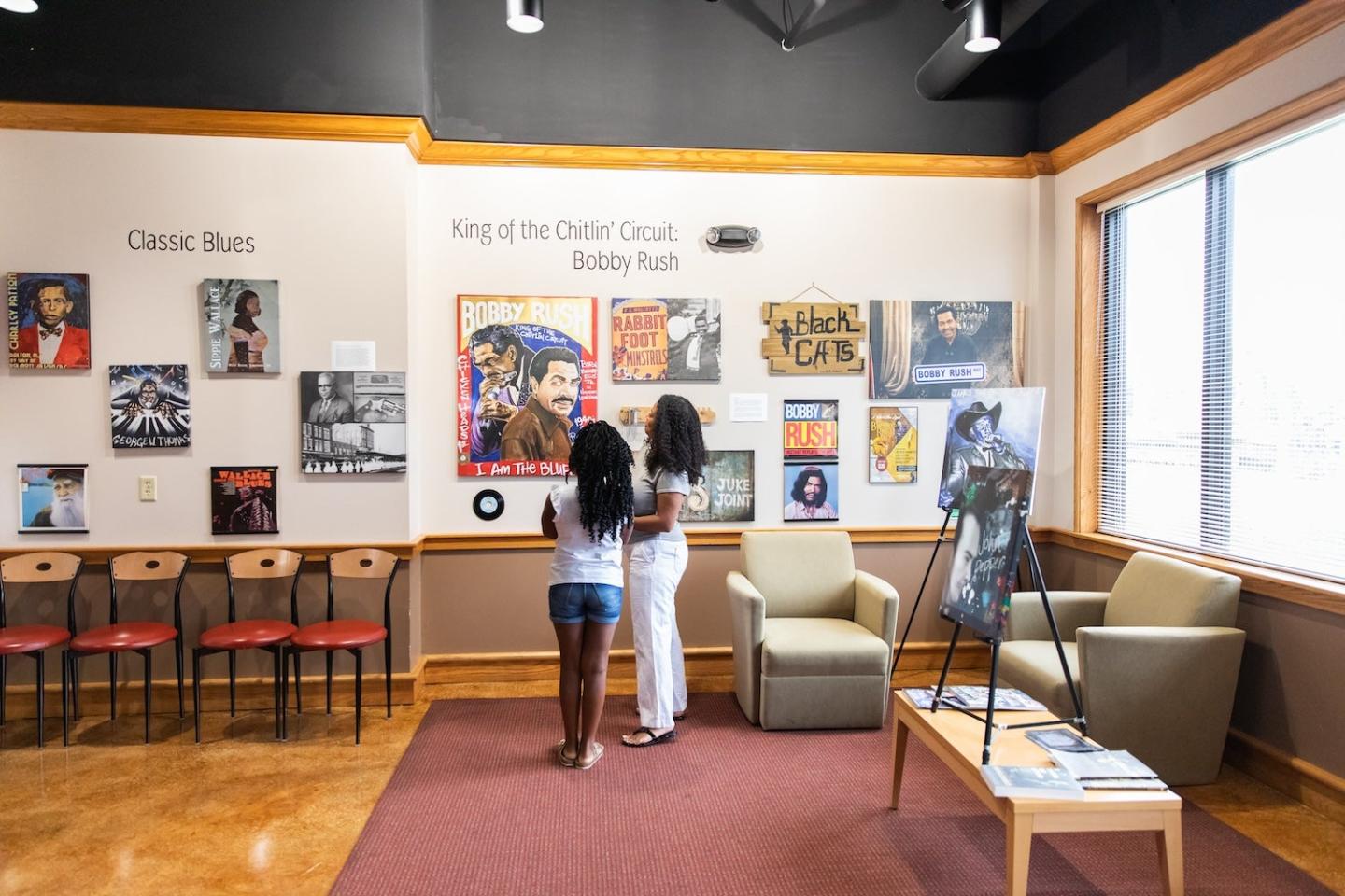 Two people in a museum exhibit with chairs and posters on the wall.