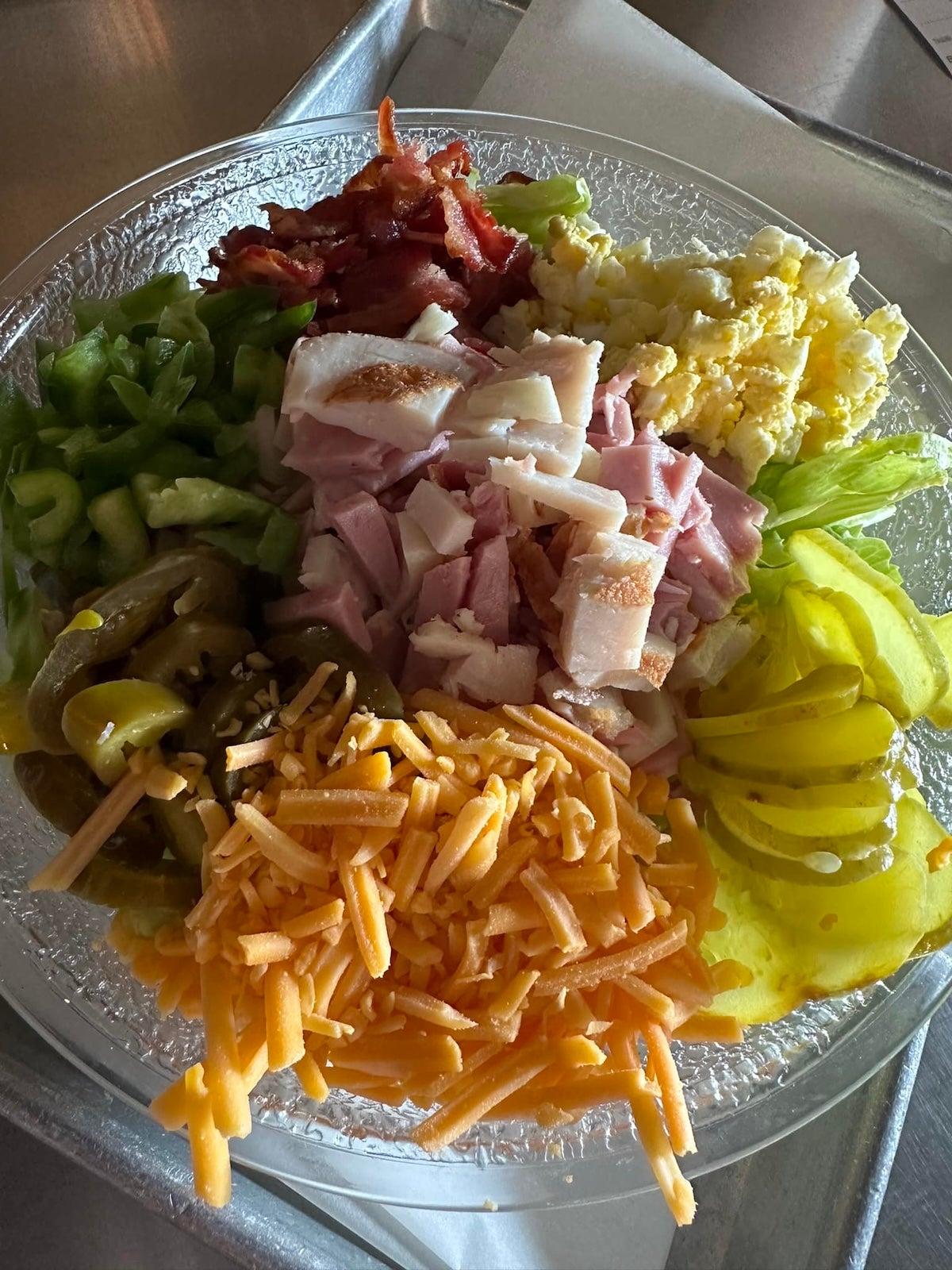 Assorted salad toppings on a clear plate: lettuce, cheese, ham, peppers, bacon.
